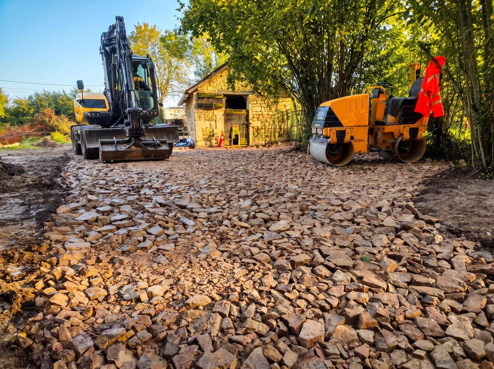 Photographie professionnelle d'un parc de matériel de terrassement en cour extérieure bien entretenue. Au premier plan, une pelle hydraulique sur pneus Mecalac jaune et noire avec bras articulé et godet frontal. À côté, un compacteur à pneus multidon visible partiellement. En arrière-plan, un camion-benne blanc et jaune aux couleurs de GRAPELOUP TP, avec benne basculante acier. Le tout stationnéé sur une surface stabilisée gravillonnée ou bétonné, hangar ou bâtiment de stockage visible flou en arrière-plan. Conditions d'éclairage naturel en extérieur, ciel dégagé. Les engins doivent être clairement identifiables et en bon état, démontrant un parc performant et bien entretenu.